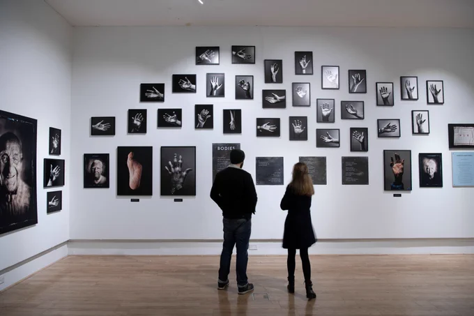 Two people exploring a white walled exhibition space. They are looking at a selection of black and white photographs of hands on the wall.