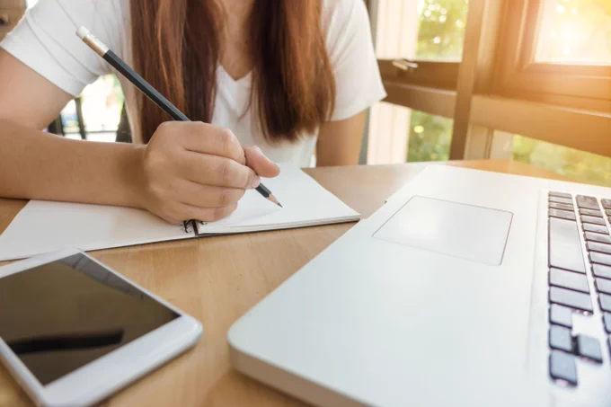 A girl sitting at a desk writing in a paper notebook with a pencil.