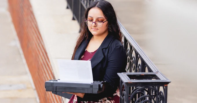A person wearing glasses is reading from a paper on a clipboard and using a laptop outdoors, standing beside a metal railing.