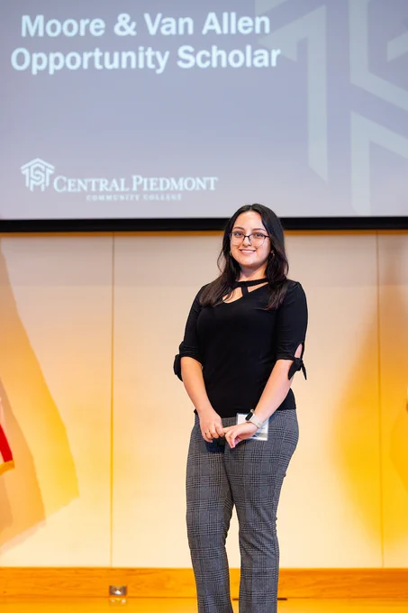 Person standing on a stage at Central Piedmont Community College, holding a microphone and smiling towards the audience.