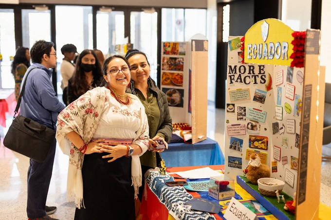 Two people smiling at a cultural exhibition booth titled "Ecuador More Facts," surrounded by banners, traditional textiles, and informative displays.