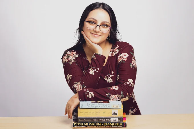 A person smiling at the camera, seated behind a stack of books including "The Norton Anthology of Contemporary Fiction" and "Popular Writing in America.