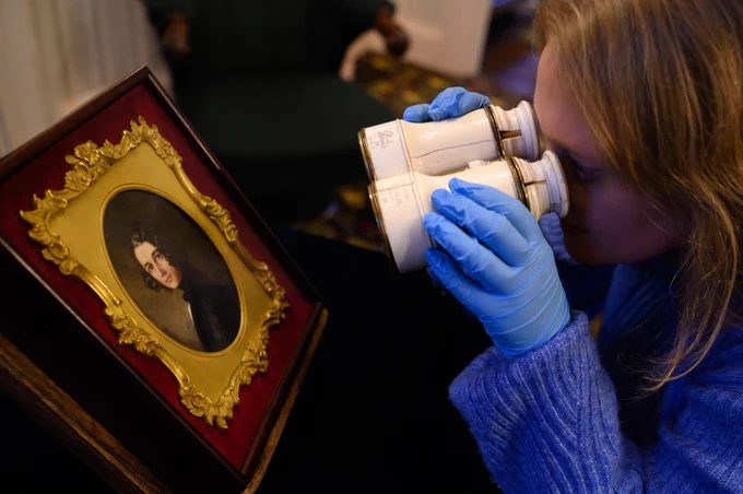 A woman, wearing blue gloves, looking through Charles Dickens’s white binoculars at “The Lost Portrait” of young Dickens.