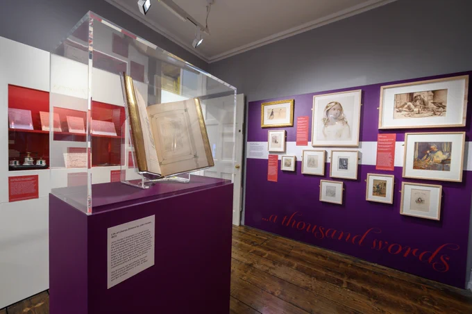 A photo of one of the exhibition rooms, with illustrations of Dickens’s characters on one wall. The “Life of Charles Dickens” by John Forster stands on a purple pedestal in the middle of the room.