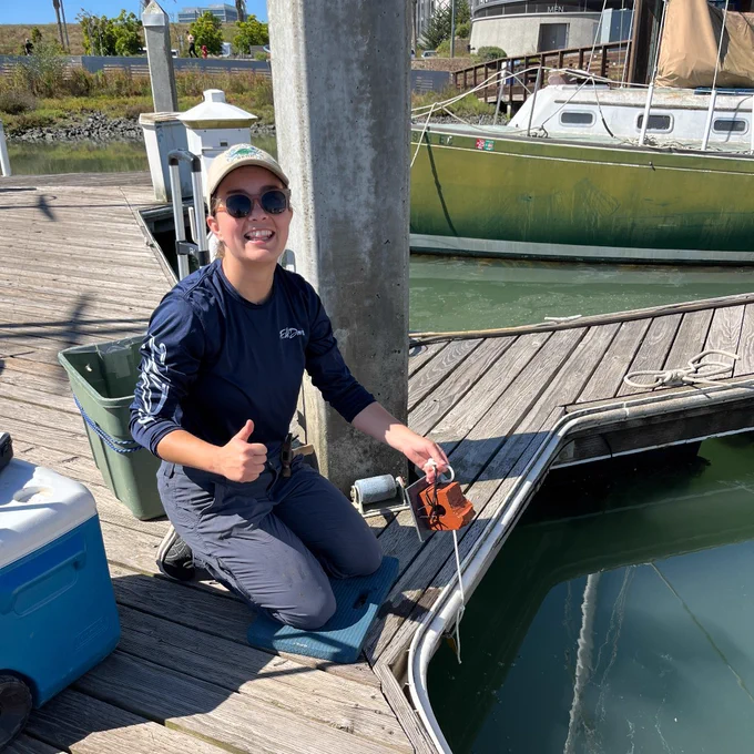 A young woman in a dark, long-sleeved shirt, blue pants, a tan hat, and sunglasses is kneeling on a dock at a marina, smiling at the camera, and giving a thumbs-up.