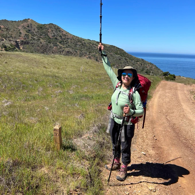 A young woman in hiking gear is standing on a dirt trail next to a large green meadow and holding a walking stick up in the air. In the background are some small mountain ranges. 