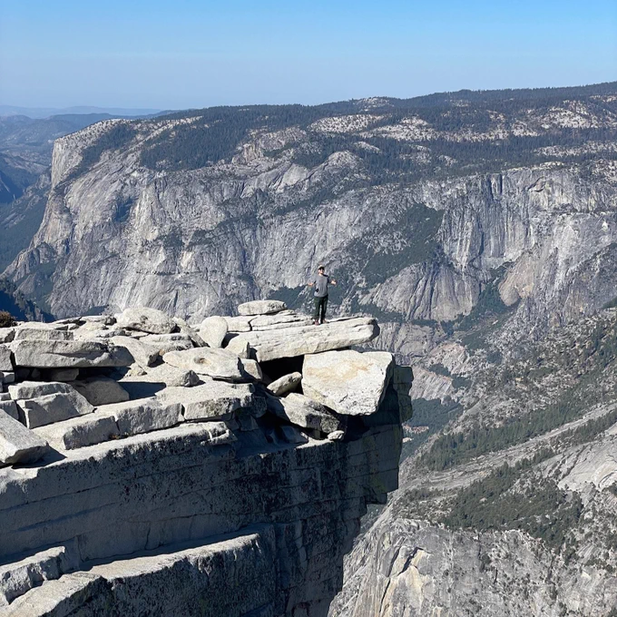 A young woman can be seen standing on a rock ledge in the distance at Yosemite National Park's Half Dome. Mountain ranges can be seen in the background. 