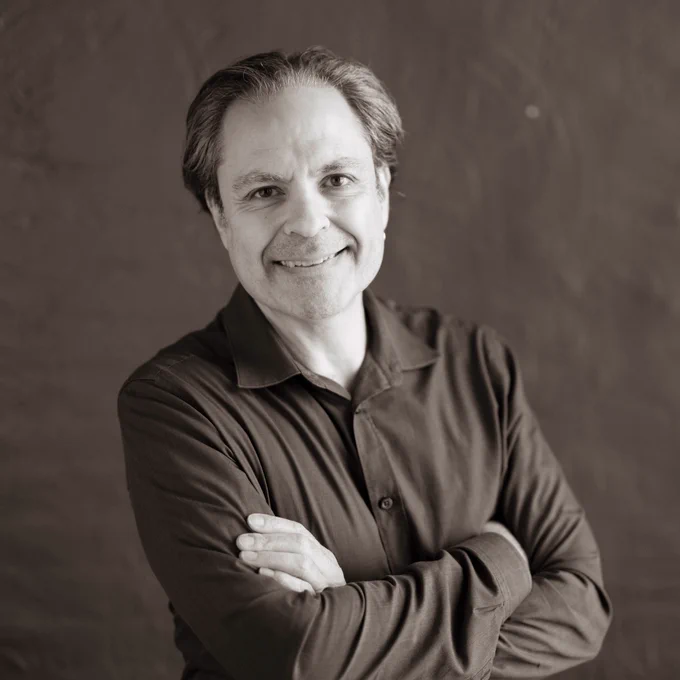 Grayscale photo of David Suisman against a studio background, arms folded and smiling at the camera.