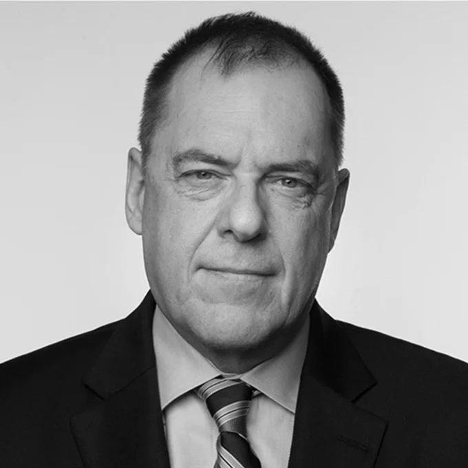 John Ibbitson wearing a suit and tie, standing against a plain white background. He has short, graying hair and a serious expression.