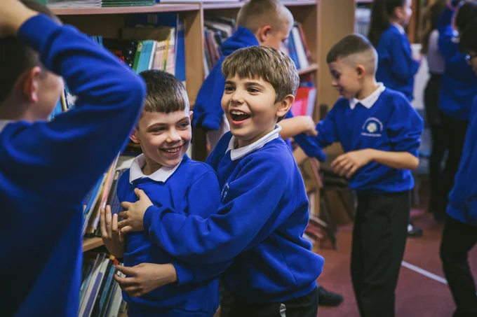 Two children wearing blue sweaters are joyfully interacting. One child, smiling broadly, reaches around and playfully hugs their friend, who is also smiling. The background features shelves packed with colourful books, and several other students in blue sweaters can be seen engaging in activities.