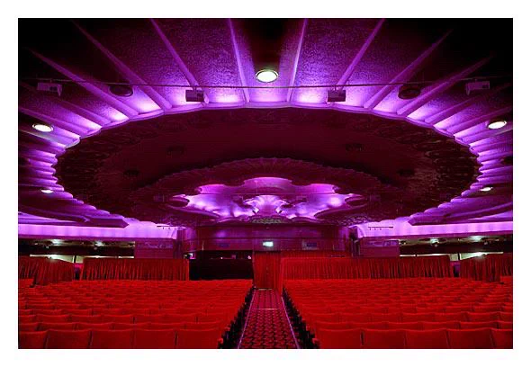 Interior of the famous Apollo Victoria Theatre with rows of red seats facing an empty stage, illuminated by purple and pink lights. This is an older design of the theatre prior to the opening of Wicked. 
