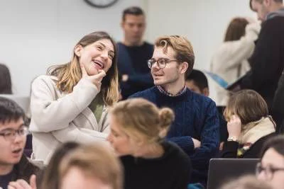 UCL Security and Crime Science students in a lively, conversational setting. A female student in a light coat and a male student in a blue sweater are smiling and talking in a crowded classroom. Around them, other students are engaged in conversation or focused on their tasks.