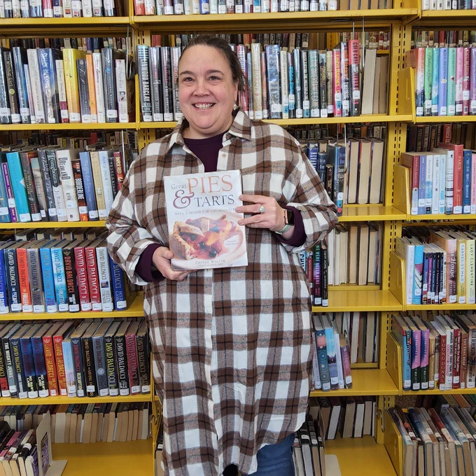 Bellefonte Library staff member stands in front of a book shelf holding a book about pie titled, Great Pies & Tarts; by Carole Walter.