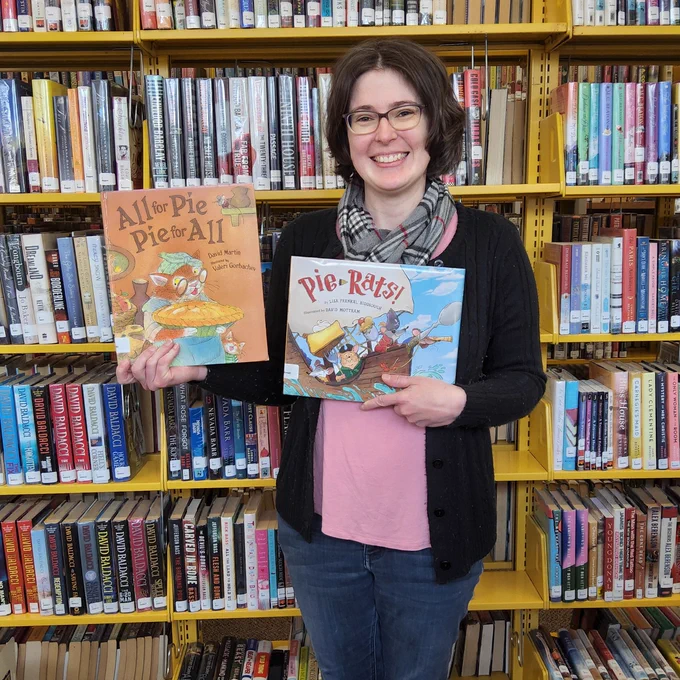 Bellefonte Library staff member standing in front of a book shelf holding two books featuring pie: All for Pie and Pie for All, by David Martin and Pie-Rats, by Lisa Frenkel Riddiough.