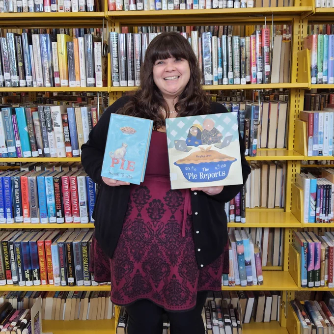 Bellefonte Library staff member standing in front of a book shelf holding two books featuring pie: Pie, by Sarah Weeks and The Pie Reports by Hayley Lowe.