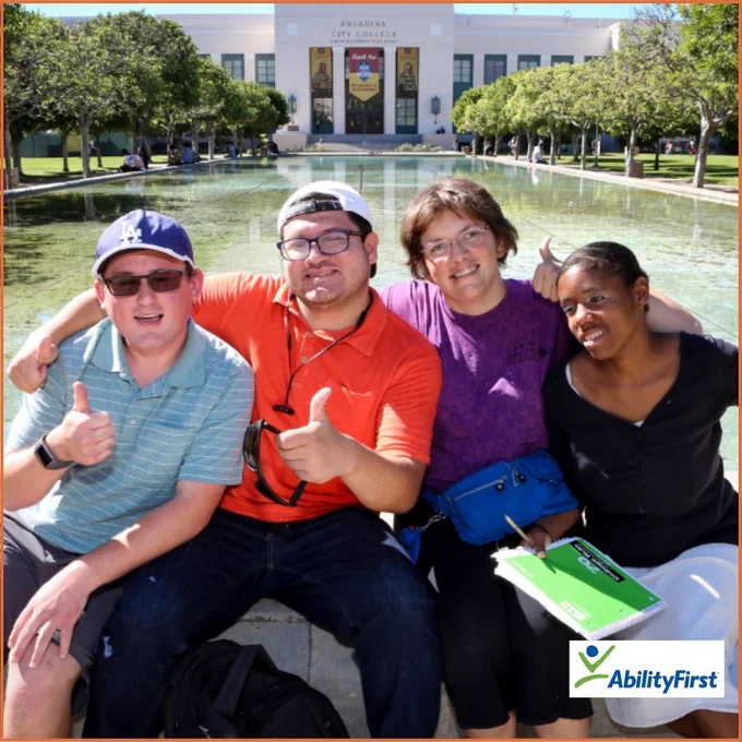 A group of 4 individuals sitting in front of a pond with a large white building lined with trees  in the background. The individuals hold their thumbs up to the camera. One woman has a notebook in her hand. The AbilityFirst logo is in the bottom-right corner.
