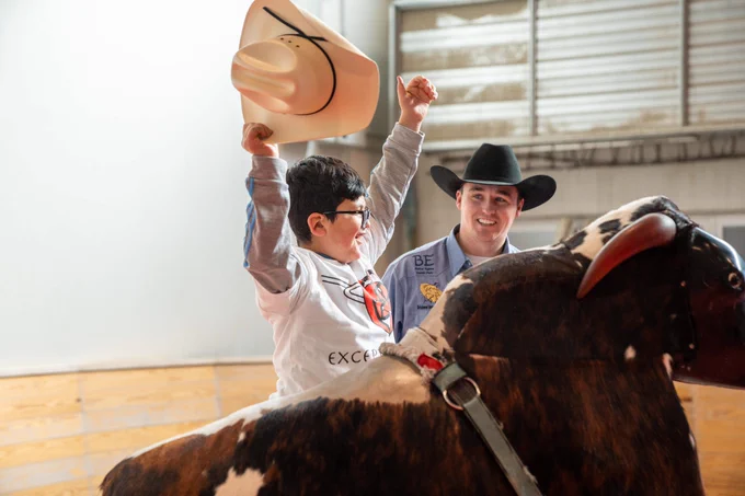 Young child smiling holding up his cowboy hat while a person stands next to them