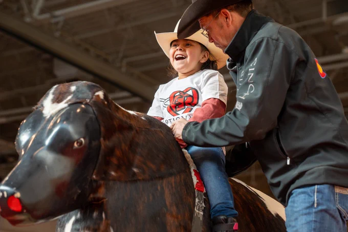 Young child being assisted on a mechanical bull by a person standing to the right
