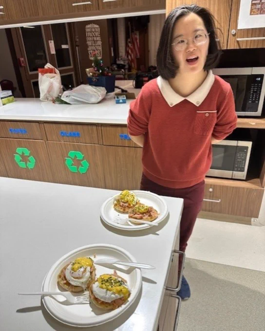 The image shows a woman wearing glasses and a red sweater with a white collar, standing in a kitchen-like environment. On the countertop in front of her are two plates of food, each containing variations of eggs Benedict, with toppings like hollandaise sauce and garnishes. Recycling bins with labels and a microwave can be seen in the background. Let me know if you’d like help crafting an accessible description for social media!