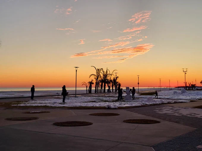 A beautifully orange sunset falls on the horizon with the Gulf of Mexico in the distance. In the foreground, people play in the snow on the beach with a snowman standing near some palm trees.