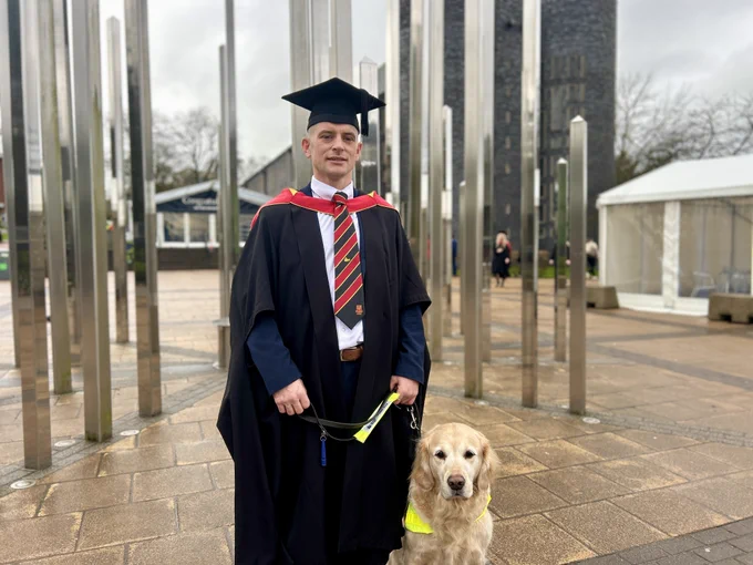 A graduate in academic regalia standing in front of a modern structure with vertical poles, accompanied by a guide dog wearing a high-visibility vest.