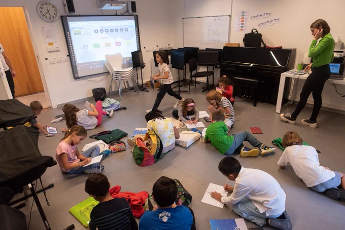 Fotografia d'uns infants fent feina al terra d'una aula. La professora, a la dreta se'ls mira.