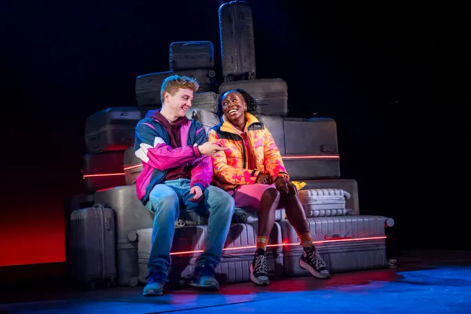 A photo showing two individuals in contemporary outfits on a darkened stage, surrounded by suitcases 