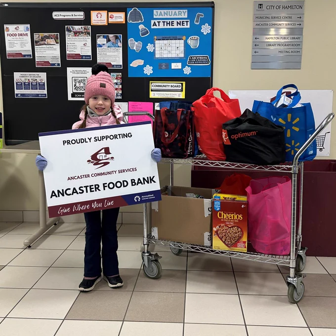 Photo of a young girl with a cart of non-perishable food donations, holding a sign that says "PROUDLY SUPPORTING ANCASTER FOOD BANK" Give Where You Live