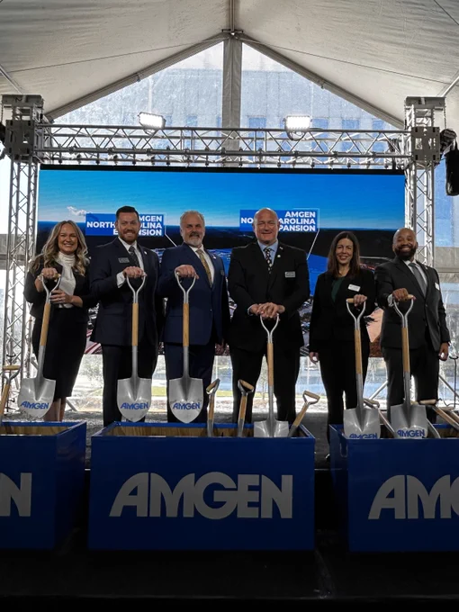 Six individuals participate in a groundbreaking ceremony under a tent, each holding a shovel with the Amgen logo on banners in front of them.