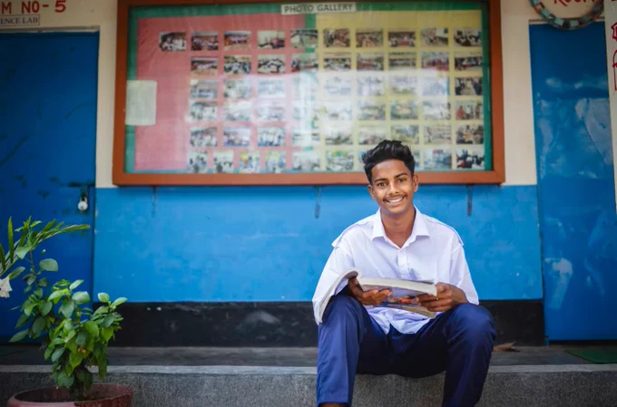 A person in a school uniform sits in front of a school photo gallery board, smiling and holding a certificate.