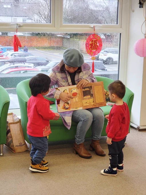 A woman wearing a blue beret and a lavender coat is sitting on a green chair reading a colorful picture book to two young children standing in front of her. Both children are wearing red tops and blue pants, attentively looking at the book. The setting appears to be indoors with large windows showing a rainy day outside. Festive decorations, including a red Chinese lantern with the word "福" (meaning fortune or happiness), hang in the window, suggesting a celebration or cultural theme. A green couch and a bag are also visible in the background.
