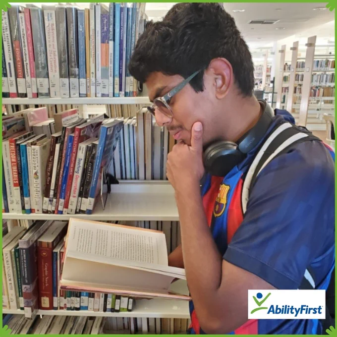 This image features a man wearing a soccer jersey reading a book standing in a library. He has his hand on his face in contemplation. The image evokes thoughtfulness and education.  The AbilityFirst logo is in the bottom-right corner.