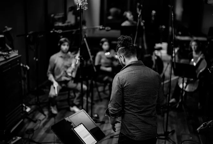 A black and white photograph from behind composer Erik Lankin showing his back and back of his head, and the front of several musicians, with their instruments, during a recording session for The Icarus Album.
Composer Erik Lankin with some of the musicians who broght Th e Icarus Album to life. 
 The whole team is Roxanne Sicard, Mattia Berrini, Pemi Paul, Silvia Buttiglione, Reuven Rothman, and Bertrand Margelidon. Devon Bate is the sound designer and the producer is Anit Ghosh. Photo: Jeff Shuh Photography