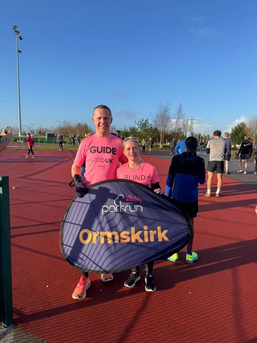 The image shows two people standing on a red running track, holding a sign that reads "Vitality parkrun Ormskirk." The person on the left is a tall man wearing a bright pink shirt with the word "GUIDE" printed on it, along with "guide running UK" and a logo below. The person on the right is a shorter woman wearing a similar bright pink shirt with the word "BLIND" printed on it. Both are smiling and appear to be participating in a running event. There are other people in the background, some walking and others standing, with a clear blue sky above.