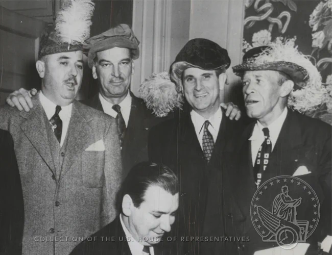 Four men in floppy hats with feathers stand behind a fifth seated man. They appear to be singing.