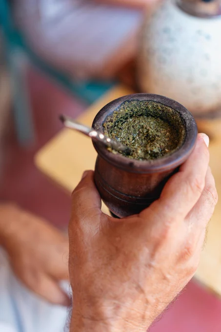 A person holding a traditional preparation of mate with crushed dried yerba mate leaves steeping in hot water in a cup made of a gourd and a specialised metal straw
