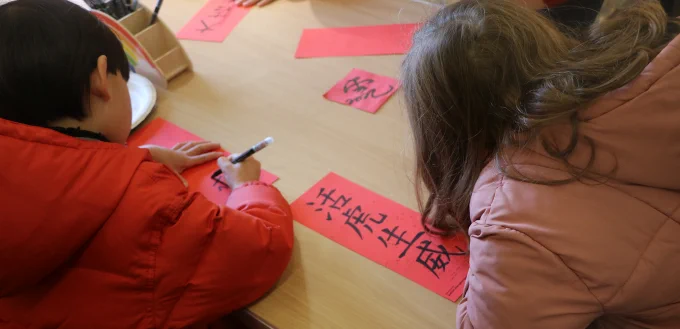 Two children are sitting at a table participating in a calligraphy activity. One child, wearing a red coat, is writing Chinese characters on a red piece of paper using a black marker. The other child, wearing a pink coat, is focused on their work. Several red papers with Chinese characters are spread across the table, indicating a festive or cultural activity for Lunar New Year.