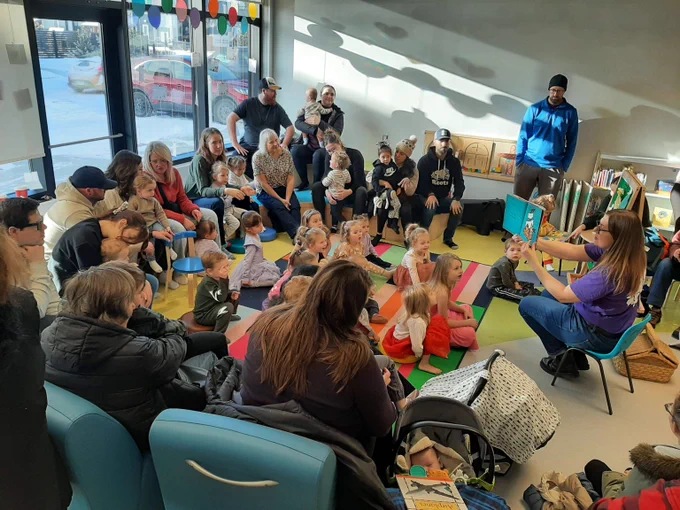 A crowd of adults with young children on their laps sit on risers that wraparound a group of children sitting on a carpet. A librarian reads them a story. 