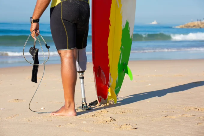 Un home amb una cama prostètica aguanta una taula de surf mirant al mar.