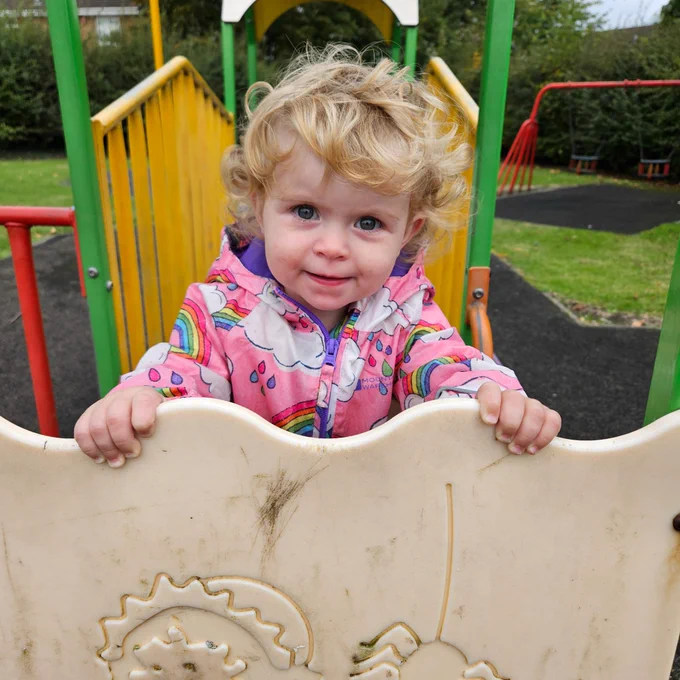 A recent photo of a smiling Annabell in a playground, wearing a pink coat with clouds and rainbows on.