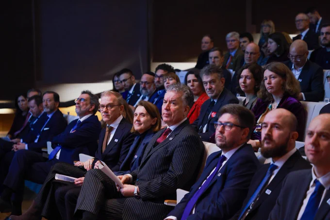 Grupa ludzi siedzi na widowni a sali konferencyjnej, wszyscy w garniturach