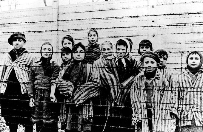A historical black and white photo of young survivors in striped uniforms standing behind a barbed wire fence at Auschwitz concentration camp.
