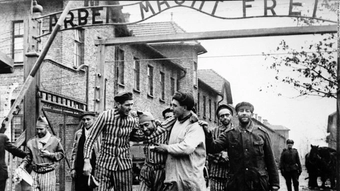 Black and white historical photo of liberated prisoners walking under the 'Arbeit Macht Frei' gate at Auschwitz Concentration Camp.
