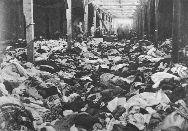 Black and white historical photo showing a large hall filled with discarded clothing, with a person visible in the background.
