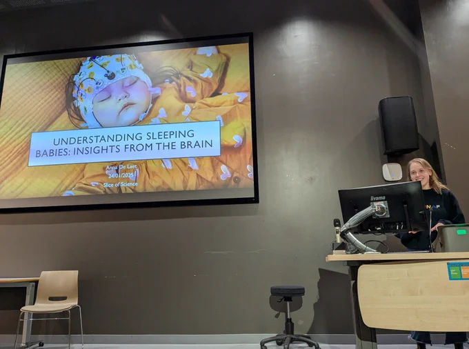 The speaker Anna De Laet, stands at a lectern in a lecture hall. On the large screen behind her, a slide shows a close-up image of a baby sleeping peacefully, wearing a patterned cap with attached sensors for brain monitoring. The baby is wrapped in a bright orange blanket with white butterfly patterns. Over the image, text reads: "Understanding Sleeping Babies: Insights from the Brain." Below it, the slide mentions "Anna De Laet, 24/01/2025, Slice of Science." 