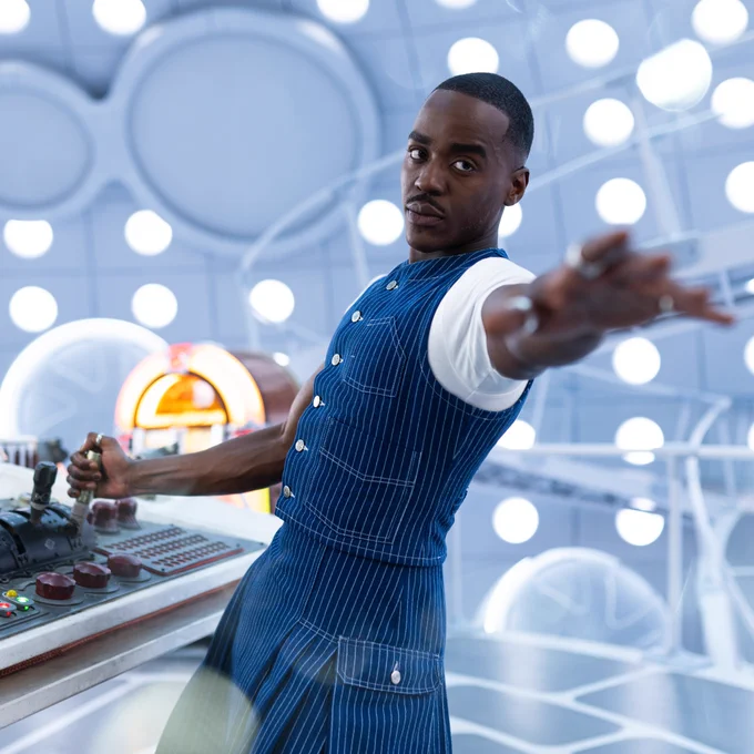 The Doctor, in a two piece navy blue pinstripe waistcoat and kilt reaches a hand towards the camera, with the other firmly on a lever on the Tardis console