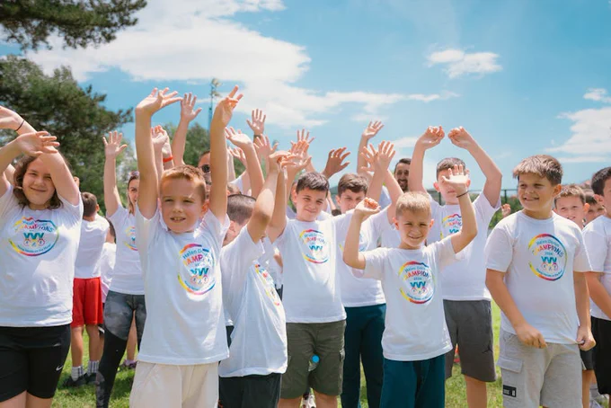 A group of cheerful children in white T-shirts raising hands in a park during summer.