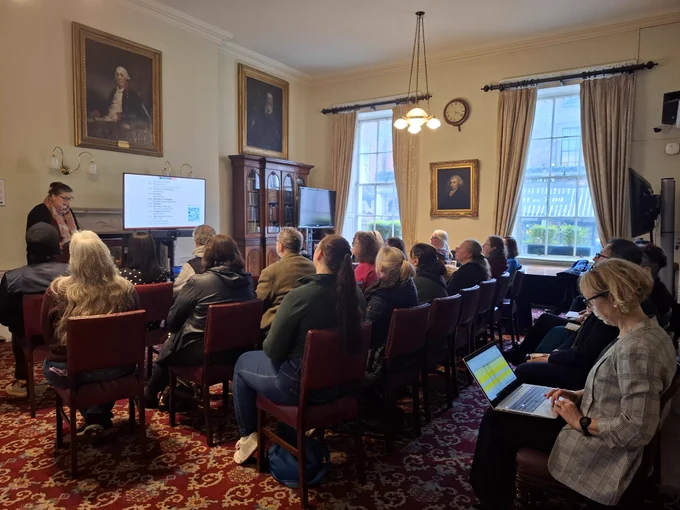 Group of people sitting and listening to a speaker with a screen that has some text on it