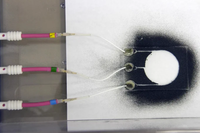 Photo of black dust particles on a white surface with white and pink wires attached to a clear rectangle of plastic or glass. The dust has scattered  in a circular pattern during an experiment for the Electrodynamic Dust Shield.
NASA
