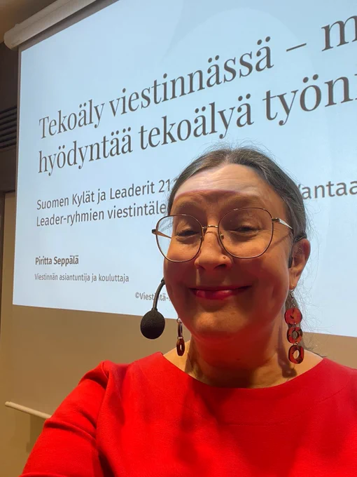 Lähikuva naisesta, joka hymyilee kameralle ja jolla on langaton mikrofoni päässä. Hänellä on punainen paita, pyöreähköt silmälasit ja riippuvat punaiset korvakorut. Taustalla on heijastettu diaesitys, jossa lukee otsikko "Tekoäly viestinnässä – miten hyödyntää tekoälyä työssä?" sekä esityksen tiedot, kuten puhujan nimi Piritta Seppälä ja tapahtuman nimi.